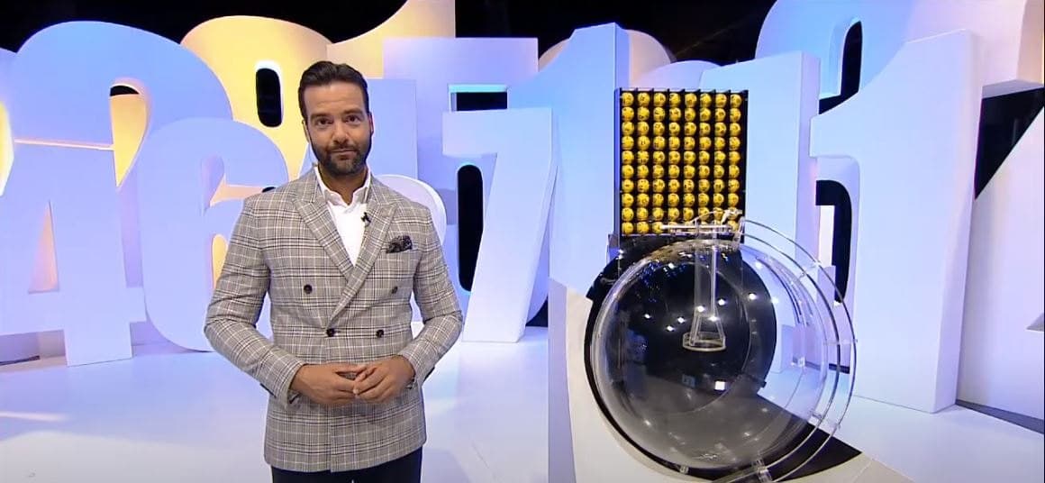 Host presenting a live televised lottery draw beside a ball-draw machine in a broadcast studio